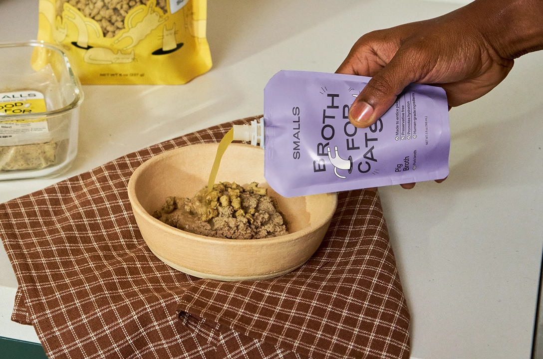 A hand pouring broth from a lilac Smalls pouch over a bowl of food on a checked cloth