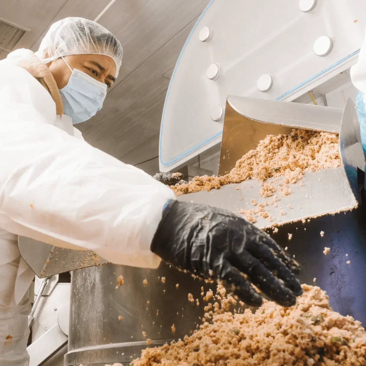 A kitchen worker in a hairnet and mask pouring cooked food from a large tub