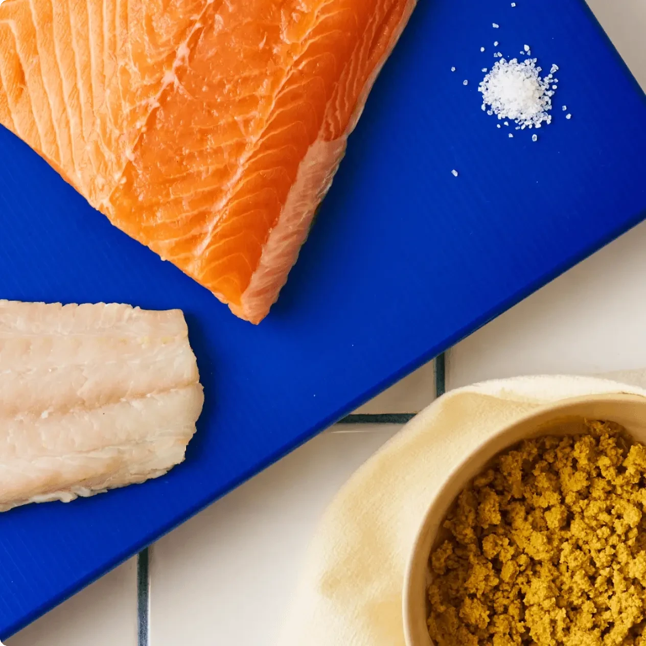 A slab of raw salmon and cod beside a bowl of kibble on a blue board