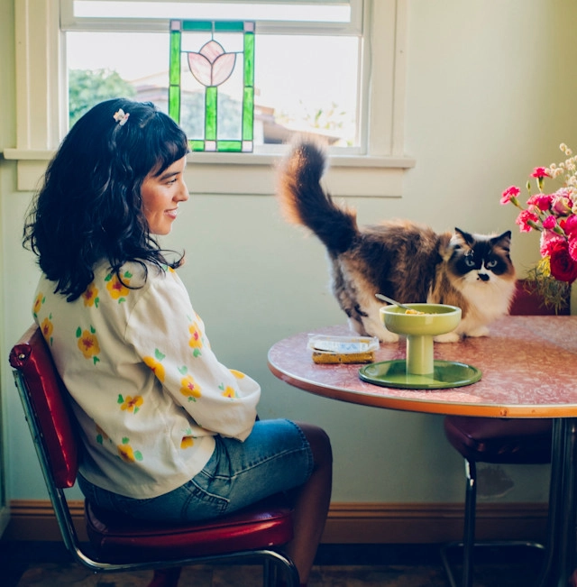 A woman at a small table by a window with a cat standing beside her