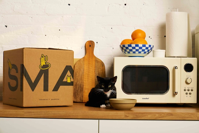 Tuxedo kitten on the counter with a bowl of food. Beside the cat is a box of Smalls, a microwave, a cutting board, and various other kitchen items.